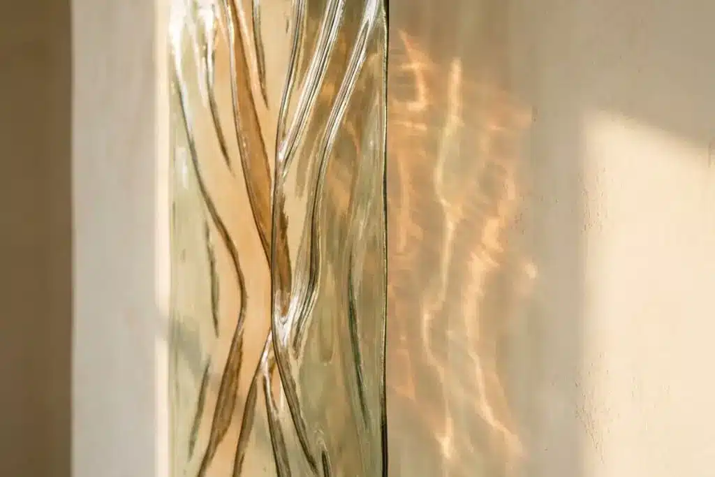 Kiln formed slumped glass panel catching afternoon daylight and casting rippled highlights on a wall.